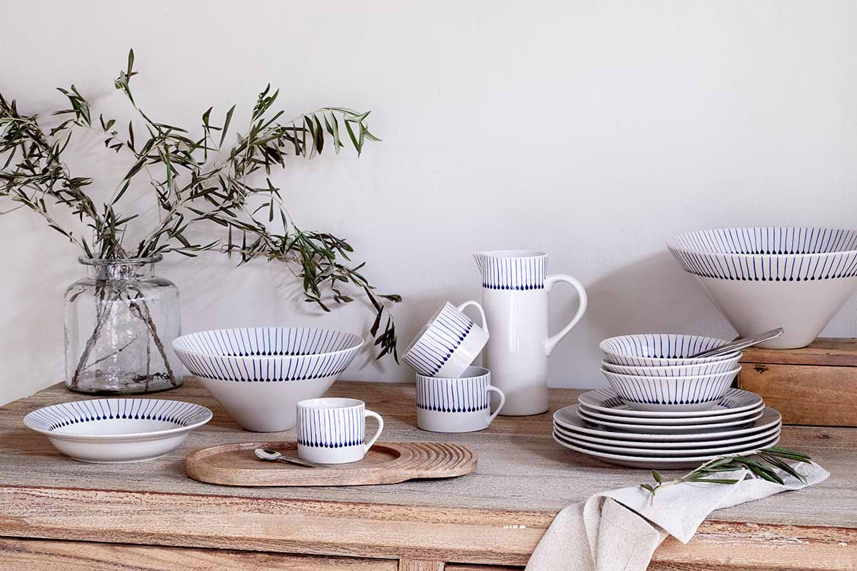 A rustic wooden table displays nkuku's handmade white clay dishes with indigo blue lines—bowls, plates, cups, and the Iba Jug - Indigo. Olive branches in a glass jar and a beige napkin by stacked plates highlight artisanal craftsmanship.