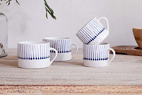 Two sets of nkuku’s Iba Mug - Indigo - Small (Set of 2) with hand-painted blue raindrop stripes rest on a rustic wooden surface, beside a wooden bowl and glass vase with greenery in the background.