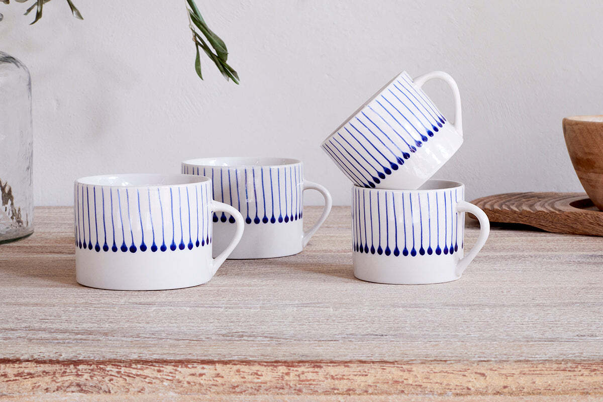 Two sets of nkuku’s Iba Mug - Indigo - Small (Set of 2) with hand-painted blue raindrop stripes rest on a rustic wooden surface, beside a wooden bowl and glass vase with greenery in the background.