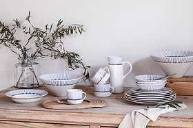 A rustic wooden table displays nkuku’s Iba Mug - Indigo - Large (Set of 2), hand-painted with thin blue stripes, alongside a pitcher, bowls, plates, leafy branches in a jar, a beige napkin, and a wooden board for a cozy minimalist look.