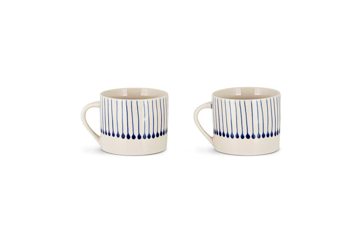 The nkuku Iba Mug - Indigo - Small (Set of 2) features white ceramic mugs with handles, decorated with vertical blue lines and indigo drop shapes for a modern, hand-painted look. Both are shown side by side on a white background.