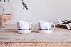 The nkuku Iba Mug - Indigo - Small (Set of 2) features two white ceramic mugs with blue vertical lines, displayed on a light wooden table with a glass jar, wooden bowl, stacked plates, napkin, and greenery in the background.