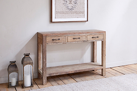 The nkuku Ibo Reclaimed Wood 3 Drawer Console Table - Natural stands by a white wall, paired with two lanterns and candles. Textured art hangs above, while wooden floors and a light rug finish the rustic hallway look.