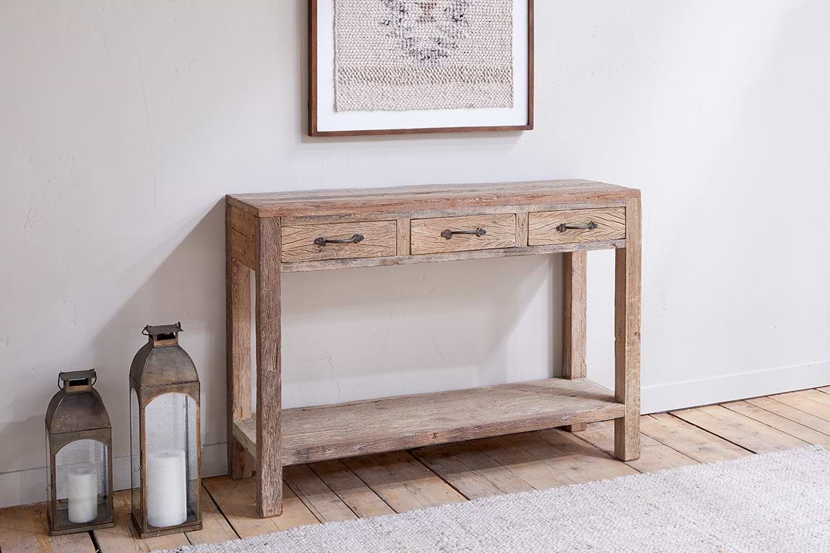 The nkuku Ibo Reclaimed Wood 3 Drawer Console Table - Natural stands by a white wall, paired with two lanterns and candles. Textured art hangs above, while wooden floors and a light rug finish the rustic hallway look.