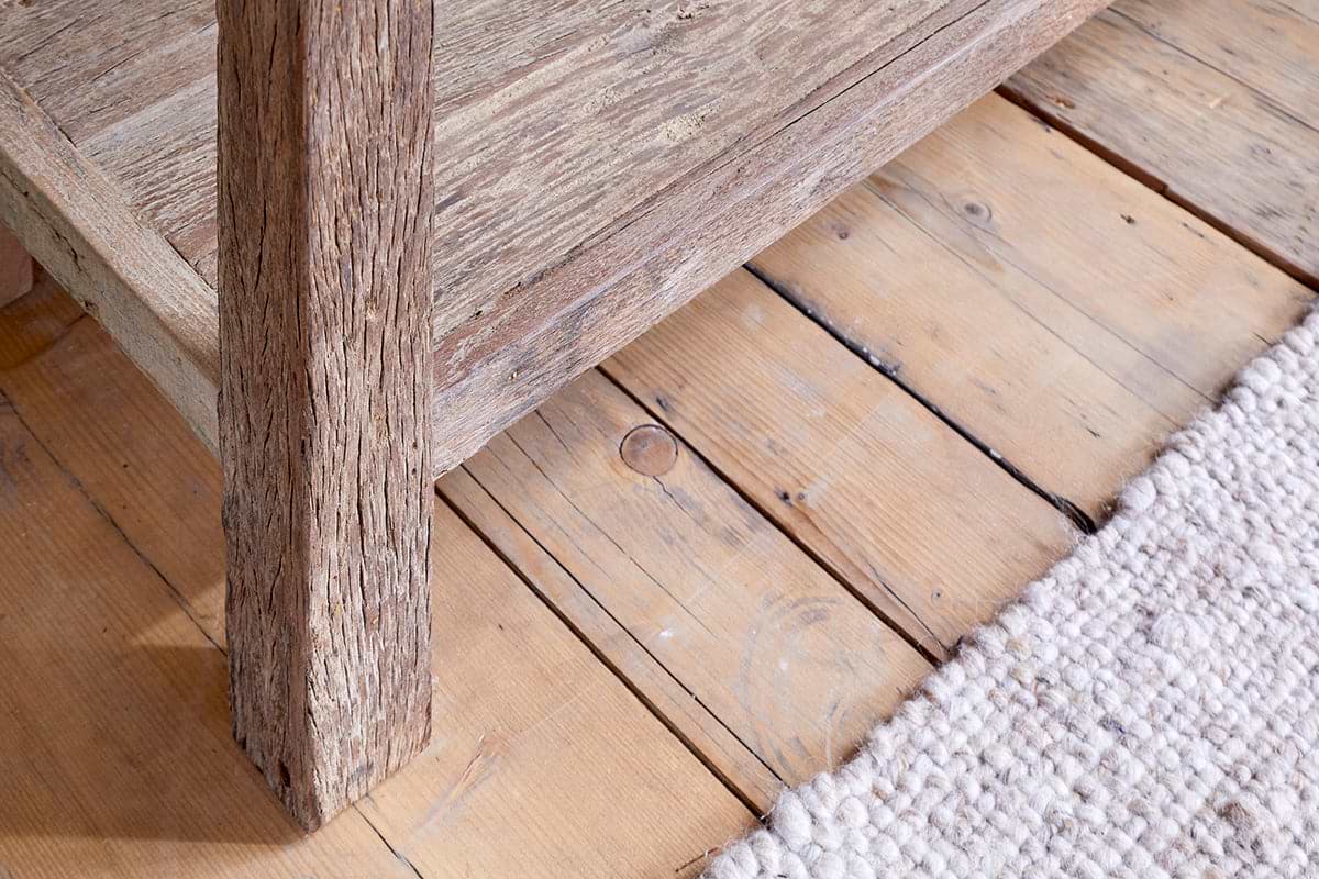 Close-up of the Ibo Reclaimed Wood 3 Drawer Console Table - Natural by nkuku, showing its rustic leg on a light wood floor. A textured cream rug highlights the natural grain and knots in both the table and flooring.