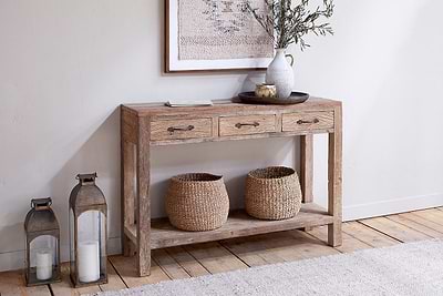 The nkuku Ibo Reclaimed Wood 3 Drawer Console Table - Natural features two baskets on its lower shelf. On top, find books, a tray, a ceramic vase with leafy branches, and a bowl. Two tall lanterns with white candles sit nearby on the floor.