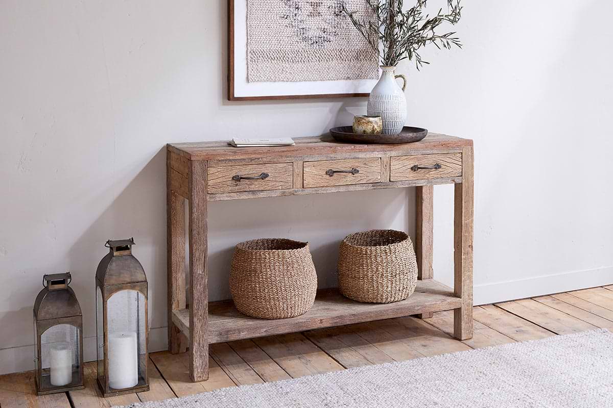 The nkuku Ibo Reclaimed Wood 3 Drawer Console Table - Natural features two baskets on its lower shelf. On top, find books, a tray, a ceramic vase with leafy branches, and a bowl. Two tall lanterns with white candles sit nearby on the floor.