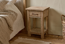 The nkuku Ibo Reclaimed Wood Bedside Table - Natural, featuring a single drawer with a metal handle and a lower shelf, sits on a wooden floor beside neutral bedding, a woven basket, and a textured rug for a cozy look.
