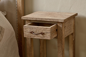 The Ibo Reclaimed Wood Bedside Table by nkuku, featuring antique hardware and an open drawer, stands beside a bed with a neutral headboard and linens.