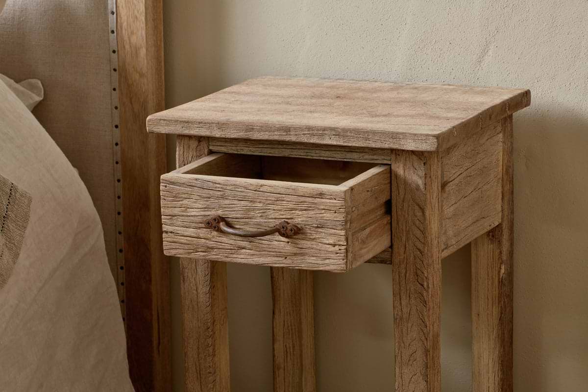 The Ibo Reclaimed Wood Bedside Table by nkuku, featuring antique hardware and an open drawer, stands beside a bed with a neutral headboard and linens.