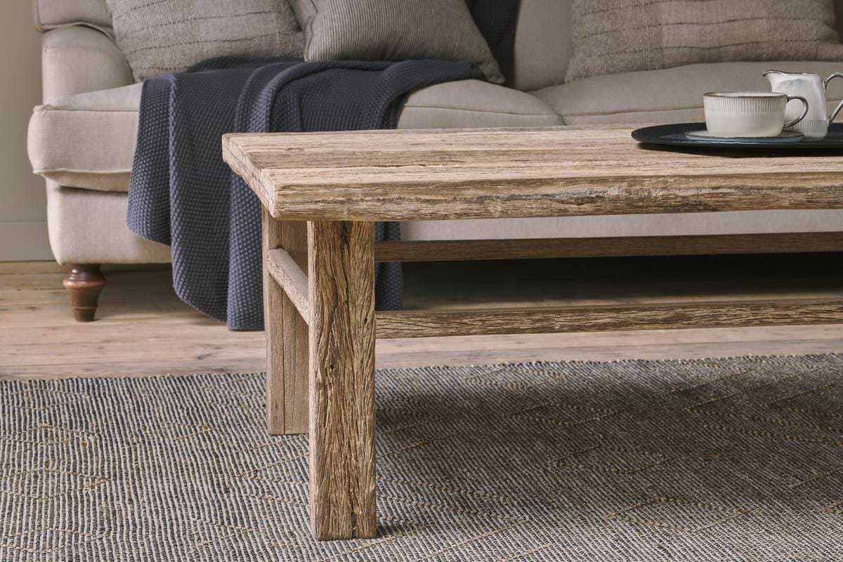 The Ibo Reclaimed Wood Coffee Table - Natural by nkuku, featuring Indian artisan craftsmanship and a textured finish, sits on a patterned rug before a beige sofa with a navy throw. A teacup and saucer are placed on the table.