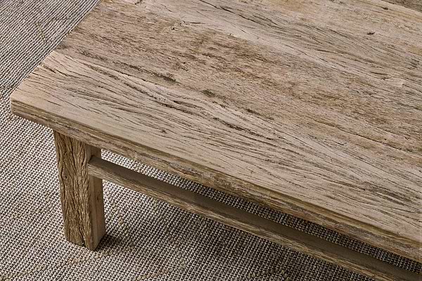 A close-up view of a rustic coffee table with a textured, weathered surface. The thick tabletop shows natural wood grain and cracks, highlighting Indian artisan craftsmanship. The table sits on a woven, tan and beige patterned rug.
