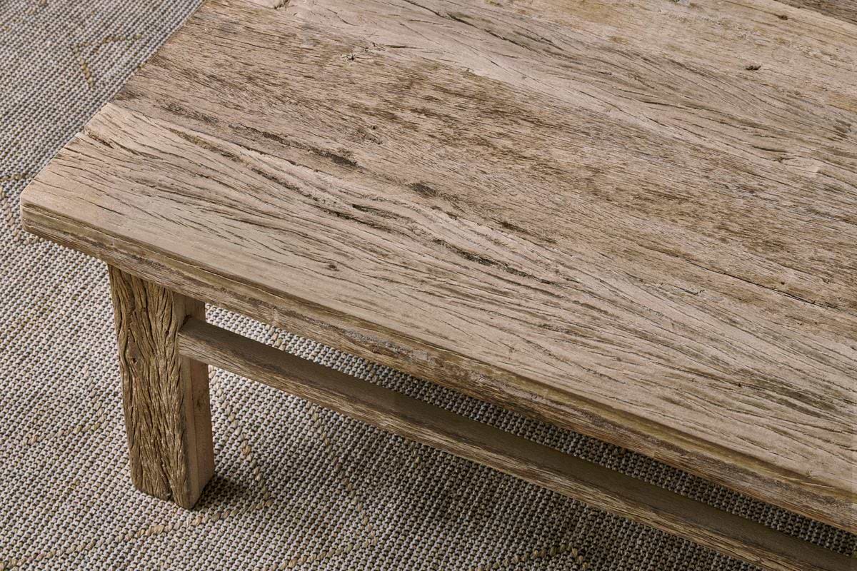 A close-up view of a rustic coffee table with a textured, weathered surface. The thick tabletop shows natural wood grain and cracks, highlighting Indian artisan craftsmanship. The table sits on a woven, tan and beige patterned rug.