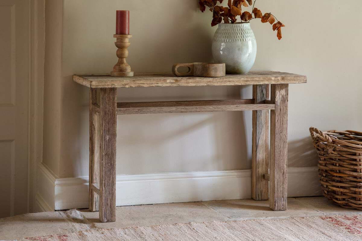 A rustic bench, reminiscent of the nkuku Ibo Reclaimed Wood Console Table - Natural, stands by a beige wall. Adorned with a ceramic vase of dried leaves, wooden candle holder, and cup; beside it lies a wicker basket and a beige-red rug.