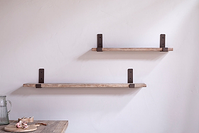 Two Ibo Reclaimed Wood & Iron Shelves by nkuku are mounted on a light wall. The lower, longer shelf sits below the shorter upper shelf. A wooden table with a small plate and pitcher is partially visible.