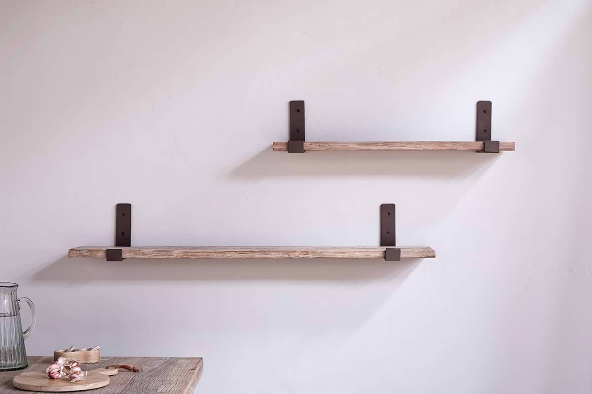 Two Ibo Reclaimed Wood & Iron Shelves by nkuku are mounted on a light wall. The lower, longer shelf sits below the shorter upper shelf. A wooden table with a small plate and pitcher is partially visible.