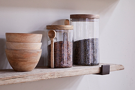Three wooden bowls are stacked beside two glass jars with wooden lids on the nkuku Ibo Reclaimed Wood & Iron Shelf – Natural. The left jar holds reddish-brown rice and a wooden spoon; the right jar contains dark lentils.