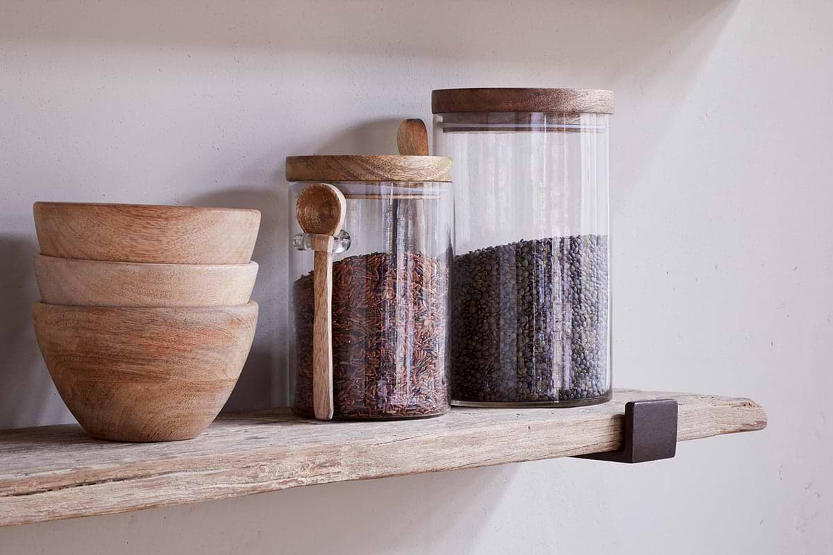 Three wooden bowls are stacked beside two glass jars with wooden lids on the nkuku Ibo Reclaimed Wood & Iron Shelf – Natural. The left jar holds reddish-brown rice and a wooden spoon; the right jar contains dark lentils.