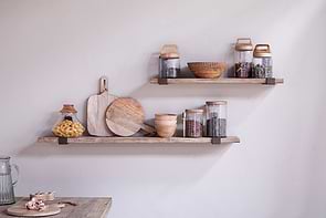 The Ibo Reclaimed Wood & Iron Shelf by nkuku, with antique brass brackets, displays jars of pasta and spices, wooden bowls, and cutting boards against a neutral wall.