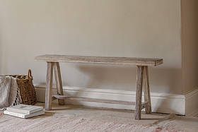 The Ibo Reclaimed Wood Long Bench - Natural by nkuku, featuring simple angled legs, rests against a beige wall. To its left is a woven basket with a folded blanket; two stacked books sit on the light, textured rug.