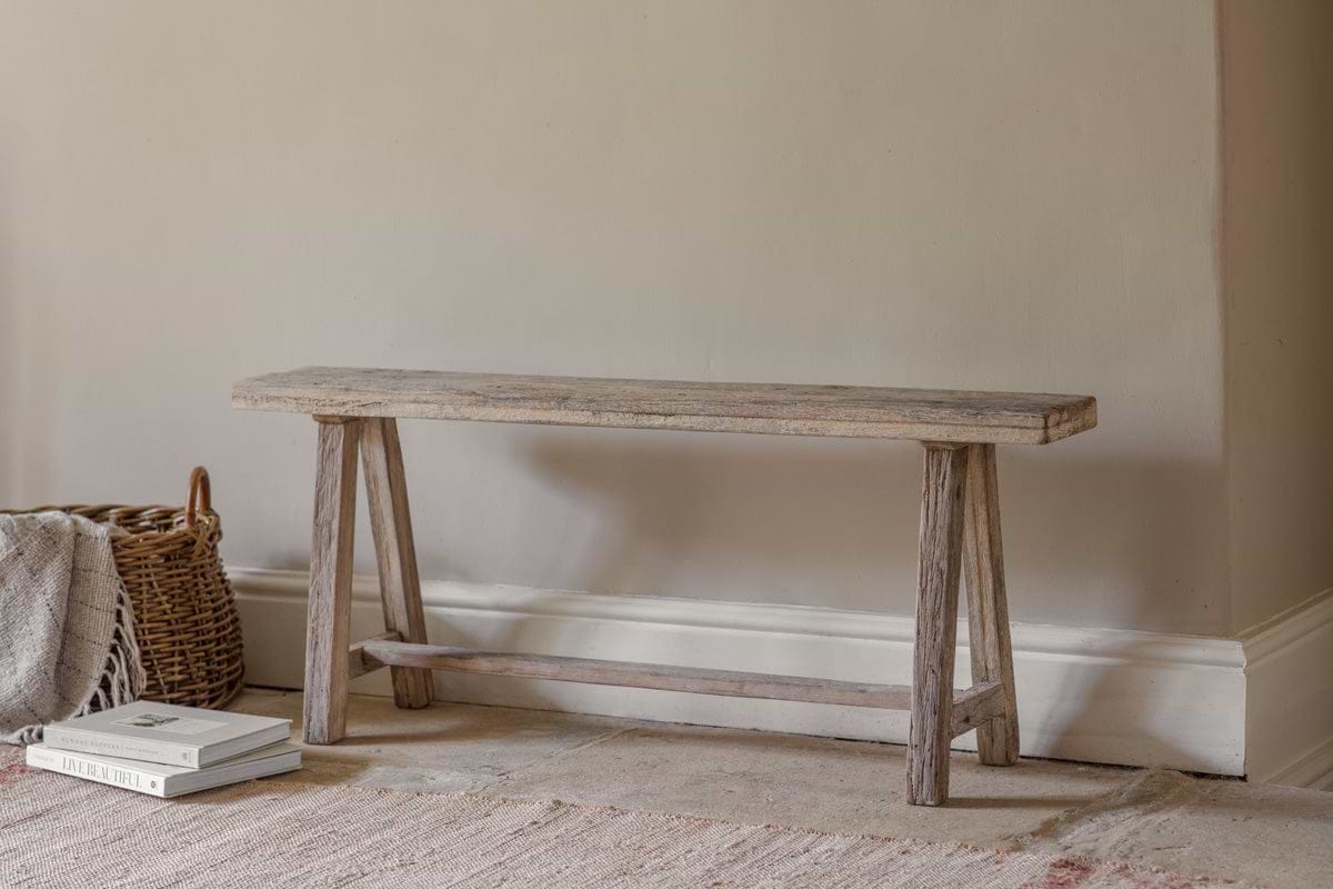 The Ibo Reclaimed Wood Long Bench - Natural by nkuku, featuring simple angled legs, rests against a beige wall. To its left is a woven basket with a folded blanket; two stacked books sit on the light, textured rug.