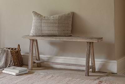 The nkuku Ibo Reclaimed Wood Long Bench - Natural, featuring angled legs, sits by a beige wall. A textured beige cushion tops the bench, while a wicker basket with a blanket and stacked books enhances the cozy, minimalist vibe.