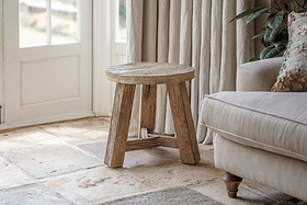 The nkuku Ibo Reclaimed Wood Round Side Table – Natural, with three legs, stands on a stone floor by a beige sofa. Sunlight streams through cream curtains onto this cozy living room accented by a leafy plant and a patterned cushion.