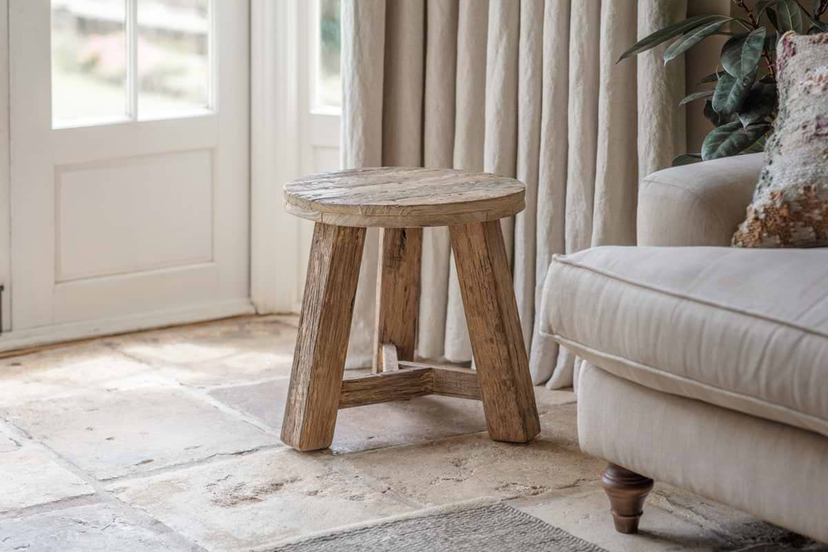 The nkuku Ibo Reclaimed Wood Round Side Table – Natural, with three legs, stands on a stone floor by a beige sofa. Sunlight streams through cream curtains onto this cozy living room accented by a leafy plant and a patterned cushion.
