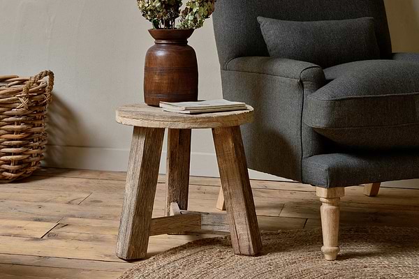 The nkuku Ibo Reclaimed Wood Round Side Table in Natural stands beside a dark gray armchair on a wooden floor, topped with a brown vase of dried flowers and two books, complemented by a woven basket and circular jute rug for a cozy scene.