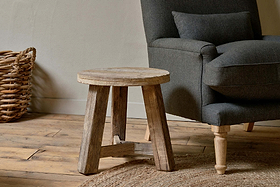 The Ibo Reclaimed Wood Round Side Table by nkuku sits on a wooden floor next to a dark gray upholstered armchair, with a woven basket and round woven rug enhancing the cozy, natural feel.