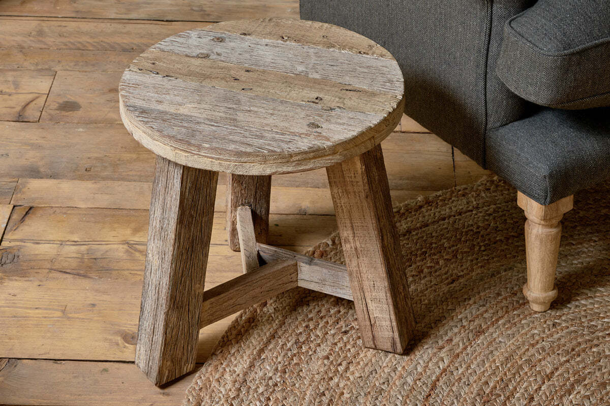 The Ibo Reclaimed Wood Round Side Table - Natural by nkuku features three sturdy legs and a weathered finish, adding cozy, Indian truck wood-inspired charm beside a dark gray sofa and woven jute rug to enhance any natural décor.