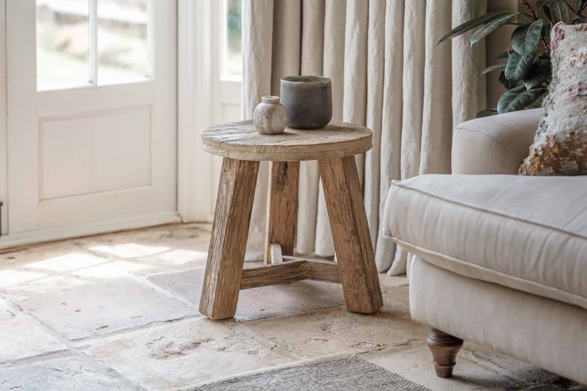 The nkuku Ibo Reclaimed Wood Round Side Table - Natural stands on a stone floor by a beige sofa and cream curtains, topped with white and gray ceramic vases as sunlight streams in through a nearby glass door.
