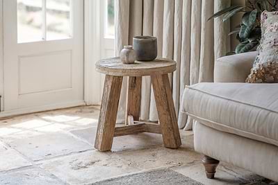 The nkuku Ibo Reclaimed Wood Round Side Table - Natural stands on a stone floor by a beige sofa and cream curtains, topped with white and gray ceramic vases as sunlight streams in through a nearby glass door.