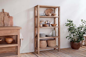 The nkuku Ibo Reclaimed Wood Shelf - Natural - Wide holds woven baskets, glass jars, ceramic bowls, plates, and mugs. A potted leafy plant stands beside it. Nearby is a wooden table with cutting boards and bowls on light wood flooring and white walls.