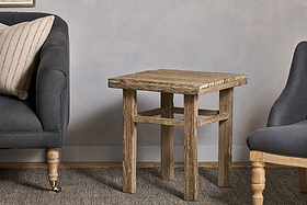 The Ibo Reclaimed Wood Side Table - Natural by nkuku, crafted with Indian artisan skill, sits between a dark upholstered sofa and armchair. Neutral tones, a striped pillow, and a textured rug complete the cozy setting.