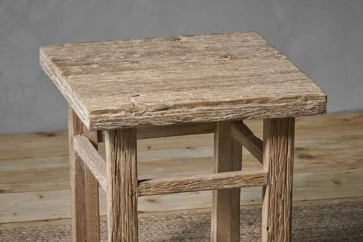 The nkuku Ibo Reclaimed Wood Side Table - Natural features a square top, four legs with horizontal supports, and a distressed finish. Handcrafted by Indian artisans, its natural wood grain and imperfections lend rustic charm to any space.