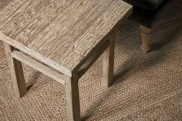 The Ibo Reclaimed Wood Side Table - Natural by nkuku, showcasing artisan craftsmanship and rich wood grain, sits beside a brown sofa on a woven natural fiber rug, adding rustic charm to the cozy space.