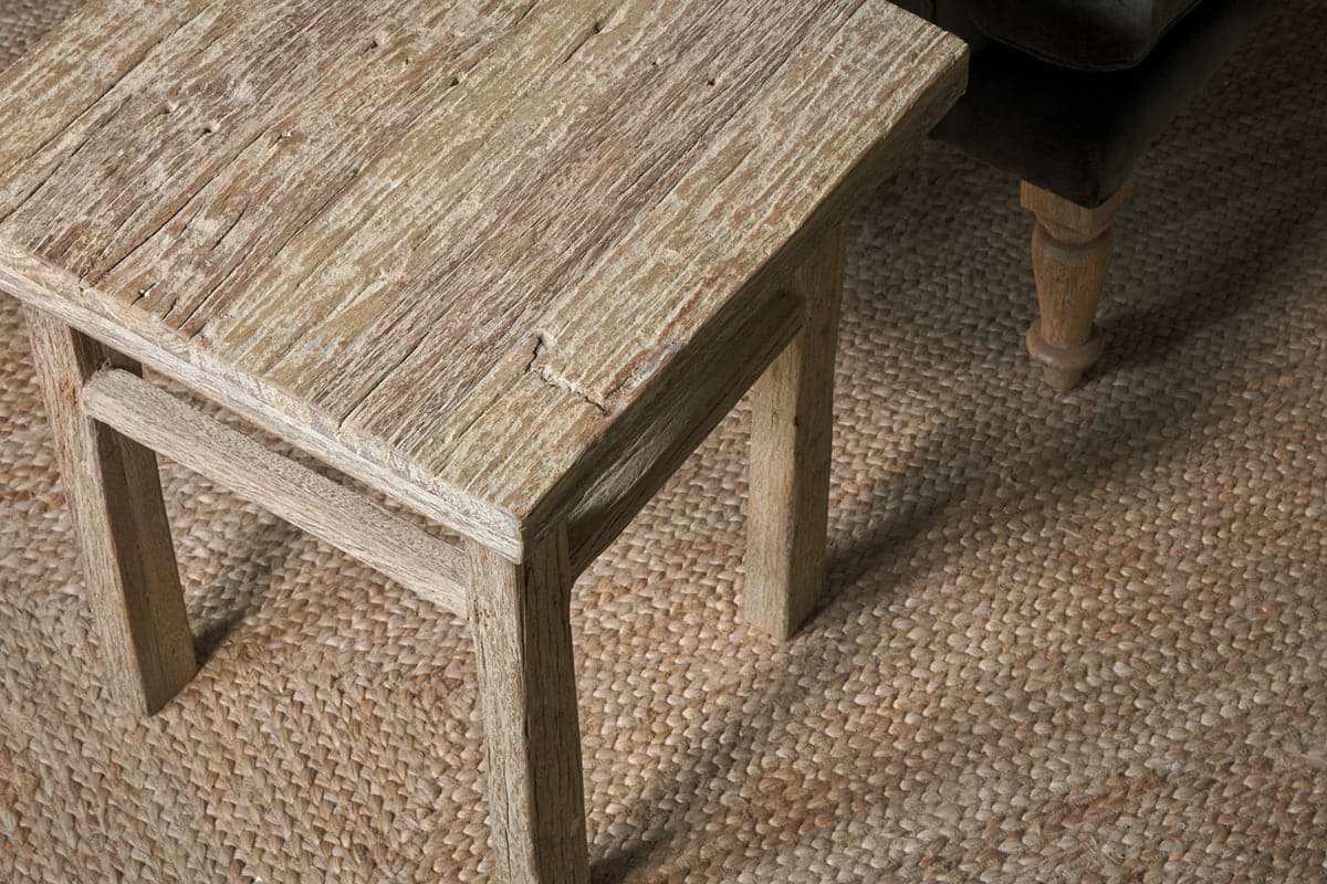 The Ibo Reclaimed Wood Side Table - Natural by nkuku, showcasing artisan craftsmanship and rich wood grain, sits beside a brown sofa on a woven natural fiber rug, adding rustic charm to the cozy space.