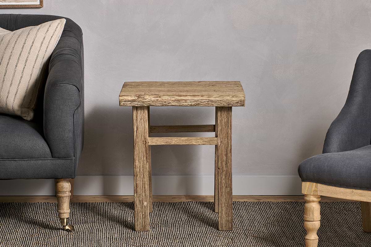 The nkuku Ibo Reclaimed Wood Side Table in Natural stands between a dark gray armchair with a striped pillow and a dark gray dining chair with wooden legs, highlighting skilled Indian craftsmanship.