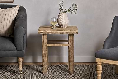 The Ibo Reclaimed Wood Side Table by nkuku, showcasing Indian artisan craftsmanship, holds a glass of white wine and a striped ceramic vase with olive branches between two upholstered chairs on a woven rug by a light gray wall.