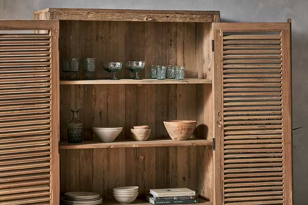 The Ibo Reclaimed Wooden Slatted Cabinet – Natural - Large by nkuku stands open against a light gray wall, revealing three shelves crafted by Indian artisans displaying glassware, bowls, cups, a carafe, and books.