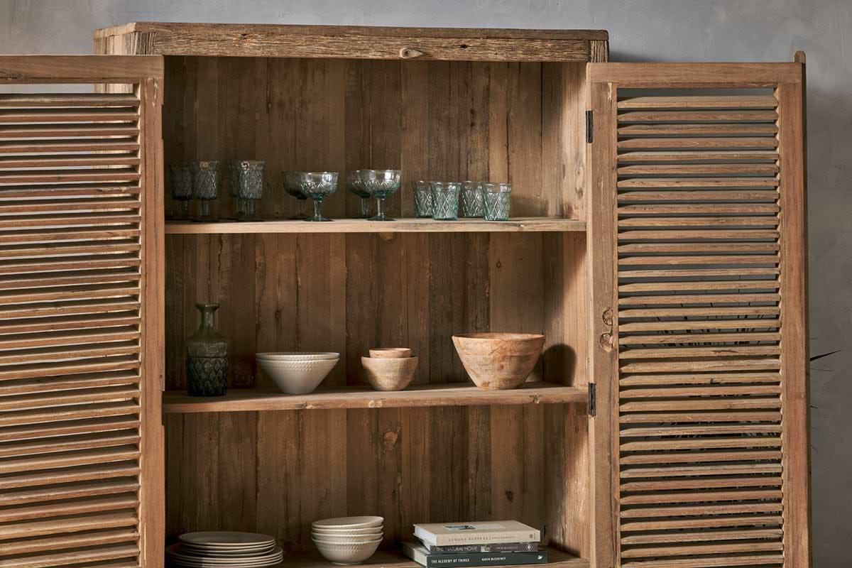 The Ibo Reclaimed Wooden Slatted Cabinet – Natural - Large by nkuku stands open against a light gray wall, revealing three shelves crafted by Indian artisans displaying glassware, bowls, cups, a carafe, and books.