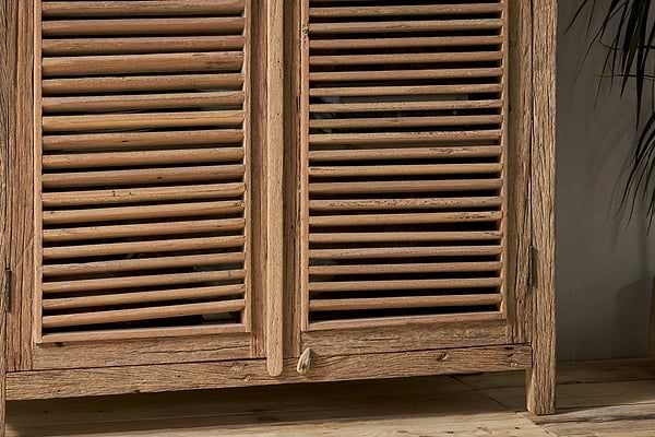 Close-up of the nkuku Ibo Reclaimed Wooden Slatted Cabinet – Natural - Large, showcasing its natural grain and knots that reflect Indian artisan craftsmanship. The rustic cabinet sits on wooden flooring next to a leafy plant on the right.