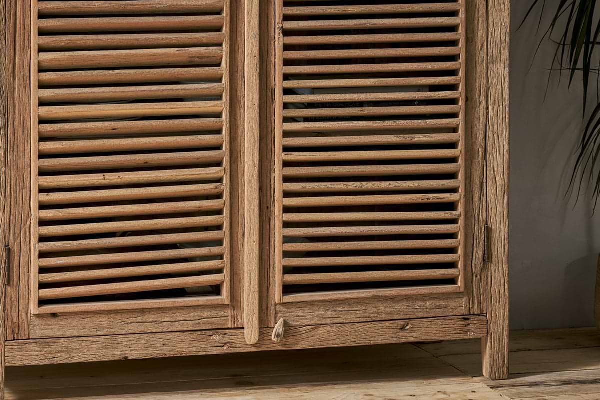 Close-up of the nkuku Ibo Reclaimed Wooden Slatted Cabinet – Natural - Large, showcasing its natural grain and knots that reflect Indian artisan craftsmanship. The rustic cabinet sits on wooden flooring next to a leafy plant on the right.