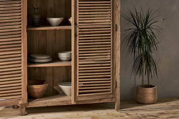 The Ibo Reclaimed Wooden Slatted Cabinet – Natural - Large by nkuku stands open, showcasing ceramic bowls and plates. Nearby, a tall potted plant with spiky leaves sits on the wooden floor against a textured gray wall.