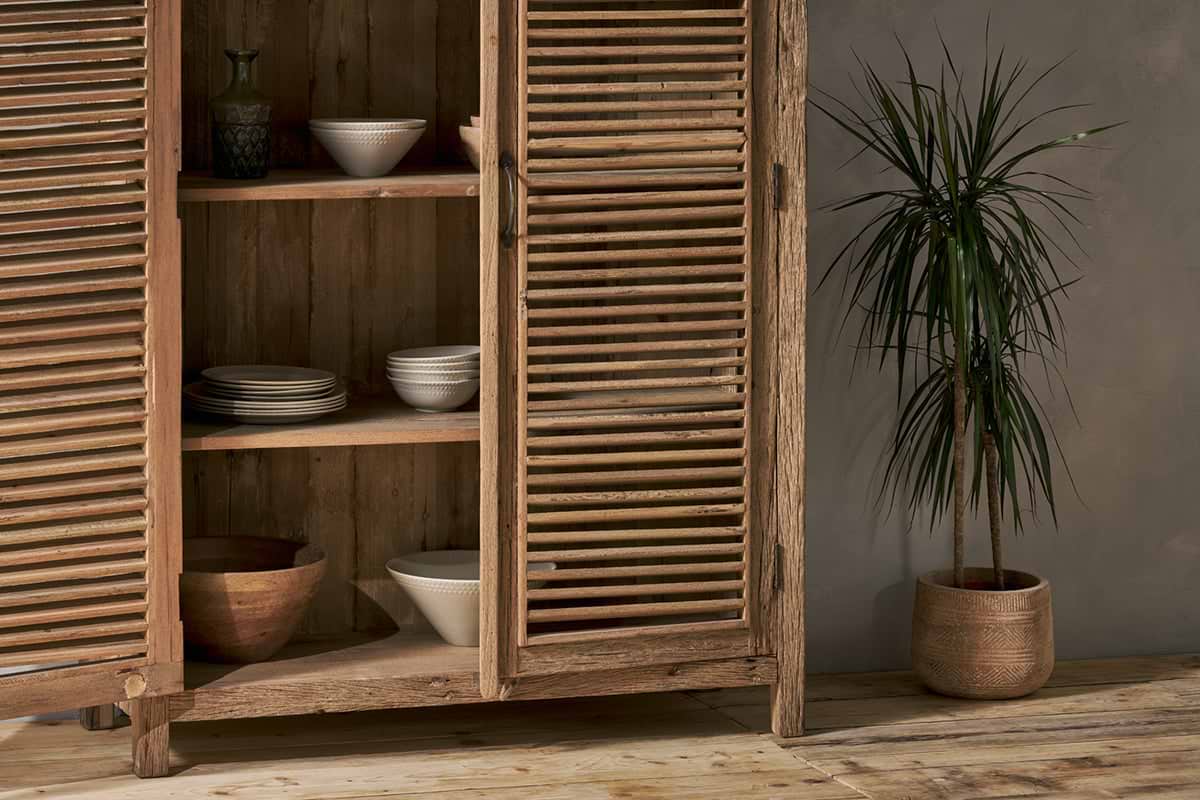The Ibo Reclaimed Wooden Slatted Cabinet – Natural - Large by nkuku stands open, showcasing ceramic bowls and plates. Nearby, a tall potted plant with spiky leaves sits on the wooden floor against a textured gray wall.