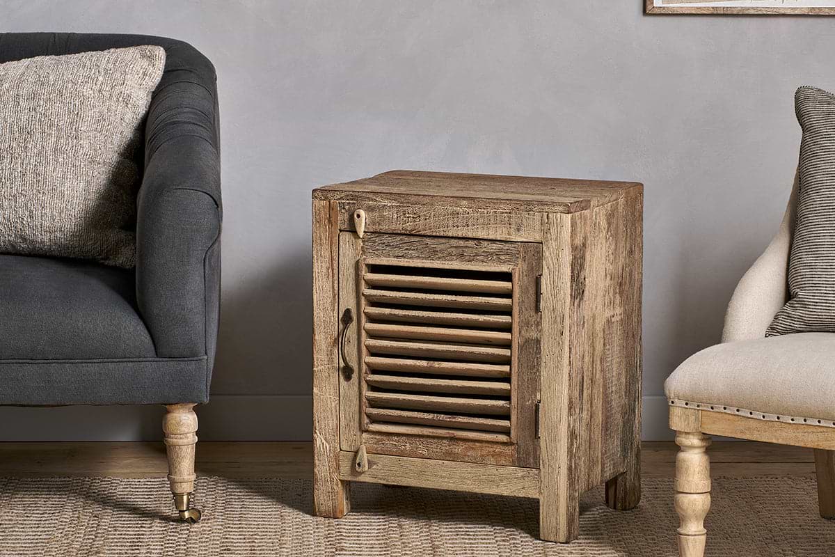The nkuku Ibo Reclaimed Wooden Slatted Cabinet – Natural - Small, handcrafted by Indian artisans, stands between a dark sofa and a beige chair with wooden legs, resting on a woven rug against a light gray textured wall.