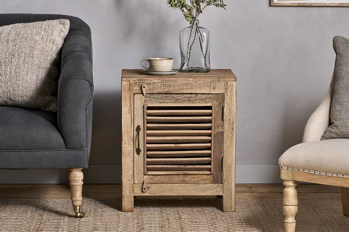 The nkuku Ibo Reclaimed Wooden Slatted Cabinet – Natural - Small, crafted by Indian artisans, stands between a dark grey armchair and a cream chair. Its slatted door with metal handle adds charm, topped with a cup, saucer, and glass vase with greenery.