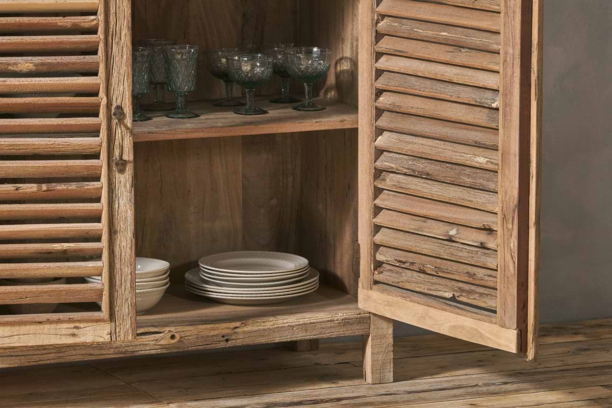 The Ibo Reclaimed Wooden Slatted Sideboard by nkuku stands open, showcasing artisan Indian craftsmanship with interior shelves holding green-tinted glass goblets and white ceramic plates above a wooden floor.