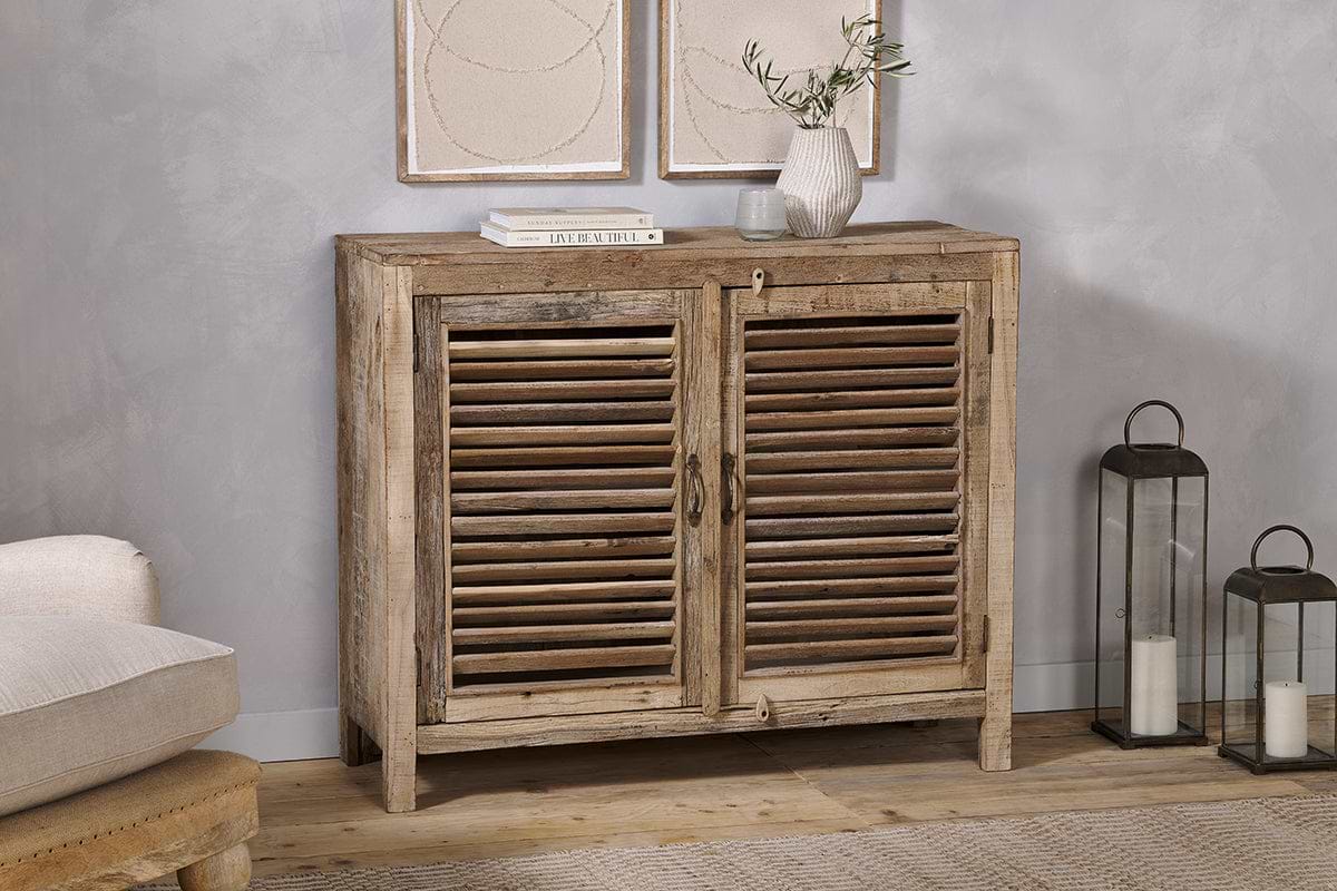 The Ibo Reclaimed Wooden Slatted Sideboard by nkuku features a natural finish and classic Indian craftsmanship. Topped with books, a vase of olive branches, and a glass, it's flanked by two metal lanterns with candles on the right.